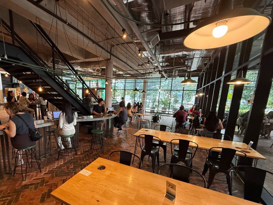 Bean Brothers at The Linc: long communal wooden tables on a herringbone brick floor in a double-height room, with steel mezzanine stairs and full-height windows